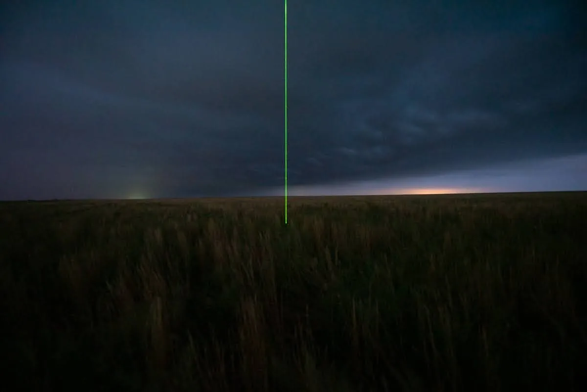 A vertical green laser beam rising from a dark Colorado prairie at dusk under a storm sky